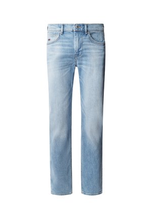 Jeune homme debout portant une veste en denim bleu clair et un jean avec des chaussures beige slip-on, regardant vers sa gauche sur un fond uni.