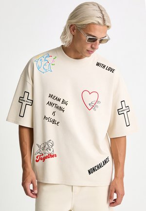 Junger Mann mit hellem Haar und Sonnenbrille, der ein beiges oversized T-Shirt mit verschiedenen bedruckten Texten und Grafiken, darunter Kreuze und ein Herz, trägt.