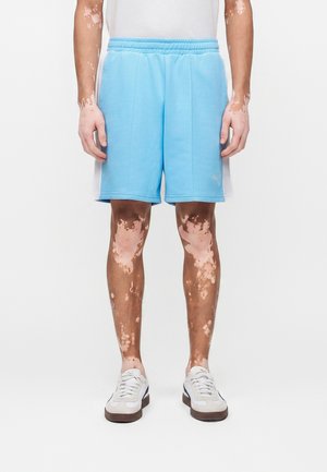 Άτομο που φοράει ανοιχτομπλε αθλητικές βερμούδες, λευκά αθλητικά παπούτσια και λευκό πουκάμισο, στέκεται με ορατά σημάδια βιτιλίγου στα χέρια και τα πόδια.