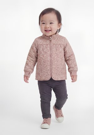 Tout-petit souriant, portant une veste matelassée rose, un pantalon foncé et des baskets roses, marchant vers l'avant sur un fond blanc.