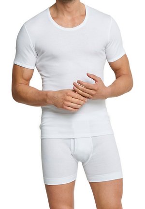 Mann in einem figurbetonten weißen kurzärmeligen Unterhemd und weißen Boxer-Shorts, der mit seiner rechten Hand seine linke Hand anpasst.