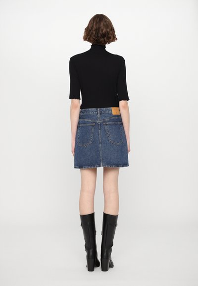 Saia de ganga com um design de cintura alta, cor azul, textura desgastada e um retalho nas costas. Usada com uma blusa preta justa e botas pretas até ao joelho.