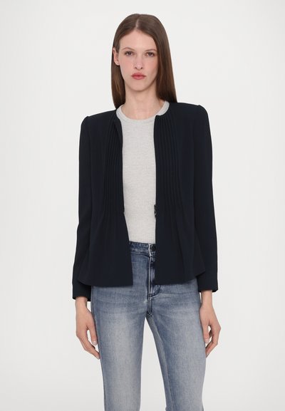 Marineblauwe geplooide blazer met een ronde halslijn, gemaakt van lichtgewicht stof. De blazer heeft een losse pasvorm en is gecombineerd met een grijze gebreide top en blauwe jeans.