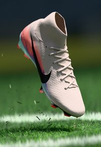 Nike football boot with a pale pink upper, black logo, and multicolored accents. Features a snug fit and raised texture for grip.