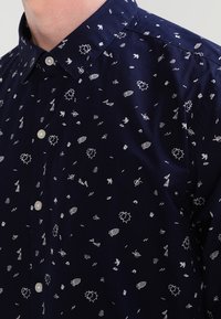 Tmavě modrá košile na zapínání s bílým vzorem tvořeným různými malými tvary, včetně šipek, listů a dekorativních prvků. Bavlněná látka, standardní límec.