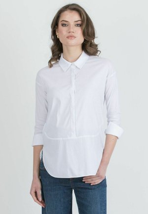 Junge Frau mit welligem braunem Haar, die goldene Creolen, ein weißes Hemd mit hochgekrempelten Ärmeln und blaue Jeans trägt, steht mit einer Hand in der Hüfte.