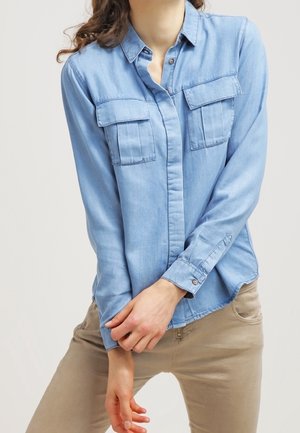 Osoba ubrana w jasnoniebieską koszulę zapinaną na guziki z kieszeniami zapinanymi na patki i beżowe spodnie, pozująca z rękami skrzyżowanymi na wysokości talii.