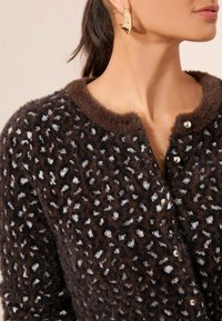 Barna bolyhos kardigán fehér és fekete pöttyös mintával, lekerekített nyakkivágással és ezüst gombos díszítéssel, arany karika fülbevalóval párosítva.