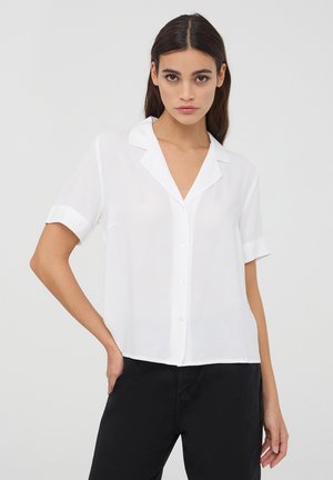 Camicia bianca a maniche corte con colletto a punta, caratterizzata da una chiusura con bottoni sul davanti e un tessuto leggero, leggermente trasparente.