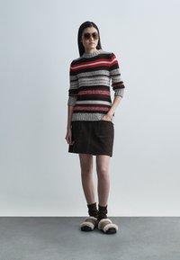 Maglione lavorato a maglia con righe rosse, nere e grigie, abbinato a una gonna in suede marrone e sandali marroni foderati in pelliccia. Accessori includono occhiali da sole.