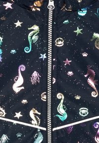 Tessuto di giubbotto nero con motivi iridescenti di cavallucci marini, stelle e corone. Presenta una zip centrale e una striscia bianca a contrasto.