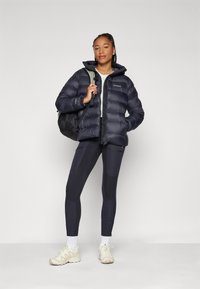 Dunkelblaues Puffer-Jacket mit horizontal gestepptem Design. Trägt es über einem hellgrauen Oberteil, kombiniert mit dunklen Leggings und cremefarbenen Sneakers.