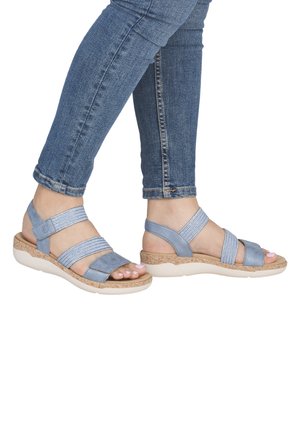 Nohy v světle modrých sandálech na pásky, doplněné o modré džíny s ohrnutými nohavicemi na bílém pozadí.