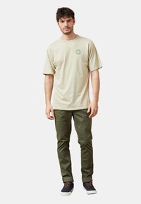 Camiseta de algodón color beige claro con mangas cortas, presenta un escote redondo y un logotipo circular. Combinada con pantalones verde oliva y zapatos oscuros.