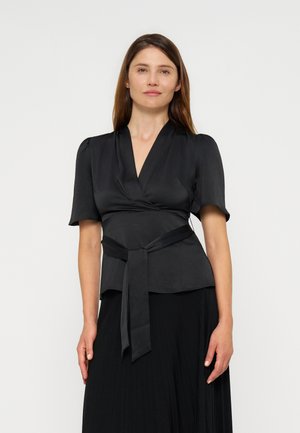 Vrouw met steil bruin haar draagt een zwarte wikkelblouse met een ceintuur in de taille en korte mouwen, staand tegen een effen lichte achtergrond.