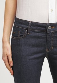 Jeans de denim azul oscuro con un corte ajustado, que presentan costuras naranjas, un bolsillo delantero y un cierre de botón. La tela tiene una textura suave.
