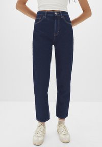 Jeans a vita alta di colore blu scuro con cuciture arancioni, taglio dritto, design a cinque tasche, tessuto denim testurizzato, abbinati a scarpe da ginnastica beige.