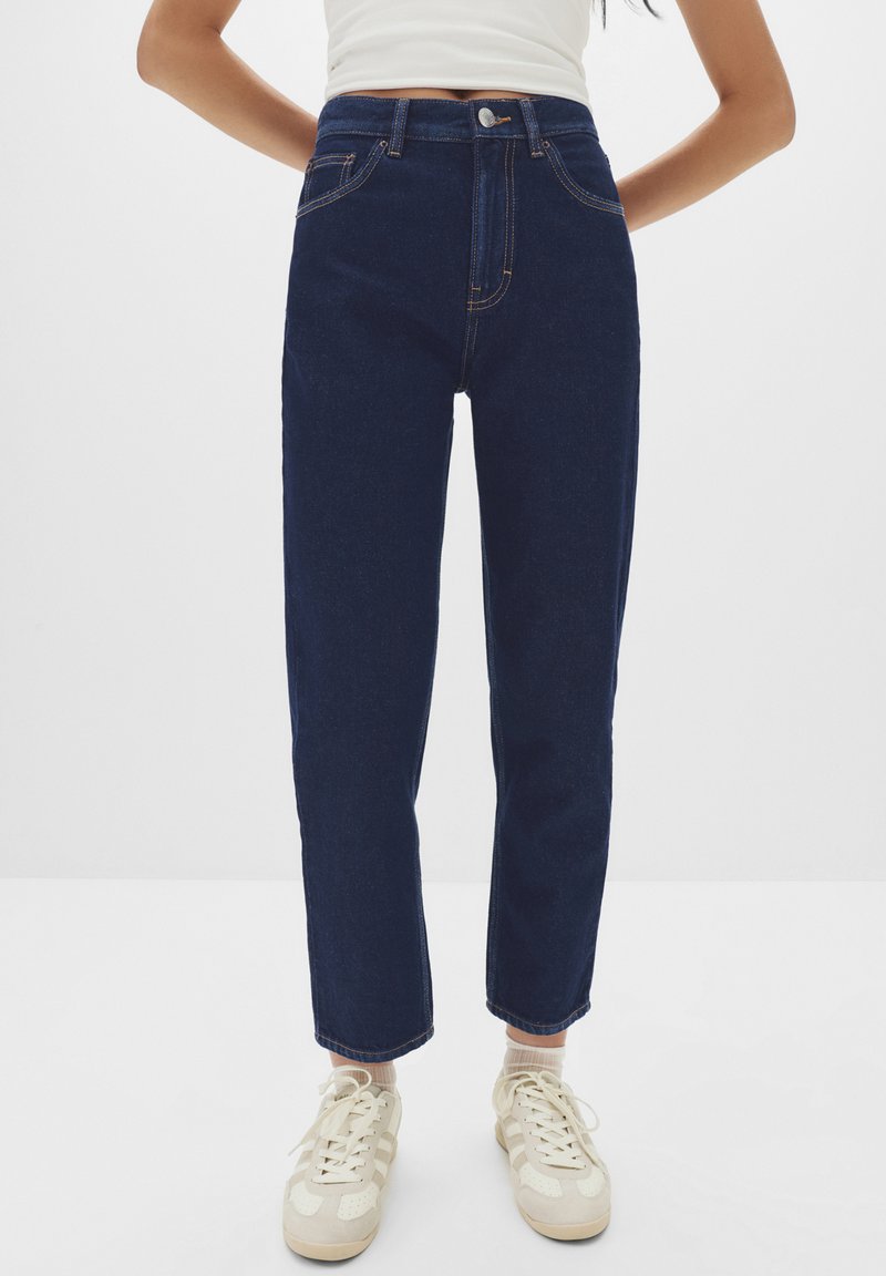 Jeans a vita alta di colore blu scuro con cuciture arancioni, taglio dritto, design a cinque tasche, tessuto denim testurizzato, abbinati a scarpe da ginnastica beige.