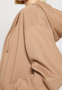 Light brown hoodie with a soft texture. Features a kangaroo pocket and drawstrings. Designed with a relaxed fit and a hood.