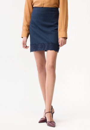 Donna con minigonna blu navy con bordo in pizzo, camicetta a maniche lunghe color senape e décolleté bordeaux con punta a punta e cinturini alla caviglia.