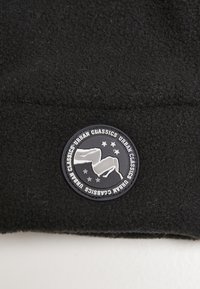Black fleece beanie featuring a round black and silver logo patch with text and graphic elements, set against a textured fabric background.