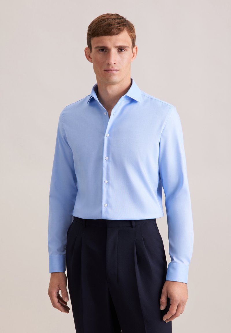 Camicia azzurro chiaro a maniche lunghe con colletto button-down e una texture liscia, abbinata a pantaloni scuri a pieghe.