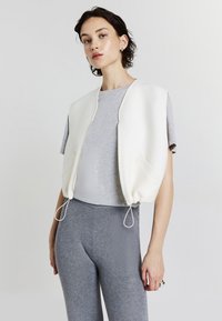 Top cropped bianco con texture liscia, chiusura con zip e orlo con cordino. Abbinato a una t-shirt grigia e leggings aderenti grigi.