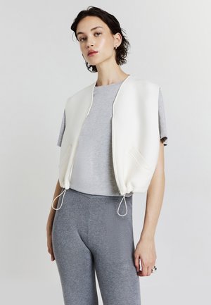 Débardeur court blanc avec une texture lisse, fermeture éclair à l'avant et ourlet à cordon. Associé à un t-shirt gris et à des leggings gris ajustés.