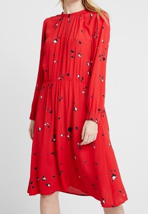 Rode midi-jurk met lange mouwen met zwart-wit bloemmotief, geplooide halslijn en taille, gedragen door een staande persoon.