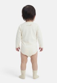 Μικρό παιδί που στέκεται με την πλάτη, φοράει εκρού ολόσωμο κορμάκι με μακριά μανίκια και το κείμενο "mon trésor" καθώς και εκρού κάλτσες, πάνω σε λευκό φόντο.