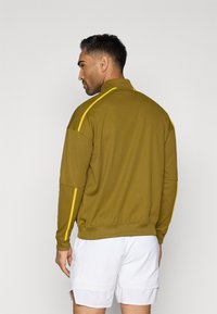 Ελαιόλαδο πράσινο φούτερ με φερμουάρ και κίτρινες ρίγες στα πλάγια, με ριγωτές μανσέτες και γιακά. Μαλακό υφή. Μοντέλο που φοράει άσπρα σορτς, με θέα από πίσω.