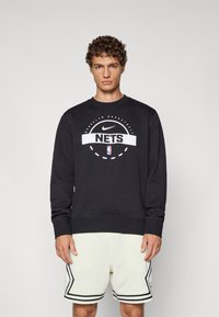 Sorte sweatshirt lavet af bomuldsblanding, med et hvidt grafisk logo og tekst. Accompanjeret af lyse, perforerede sportsshorts med sorte accenter.