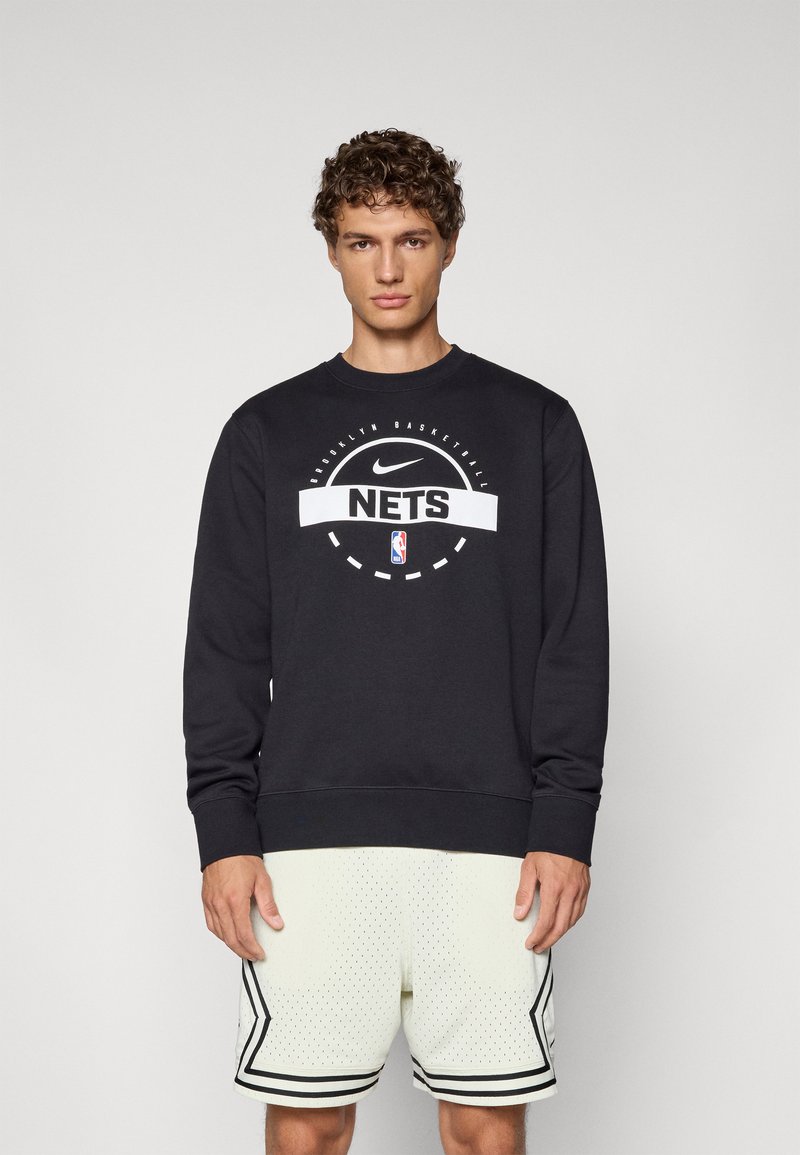 Sudadera negra hecha de una mezcla de algodón, con un logotipo gráfico blanco y texto. Acompañada de pantalones cortos deportivos perforados de color claro con acentos negros.