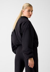 Veste noire oversize avec poignets et ourlet élastiques. Dotée d'un col montant et d'un tissu lisse légèrement brillant. Modèle vu de dos.