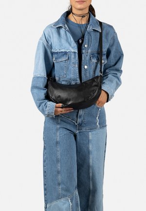 Black leather shoulder bag with a crescent shape, smooth texture, and a thin strap, held by an individual wearing a denim outfit.
