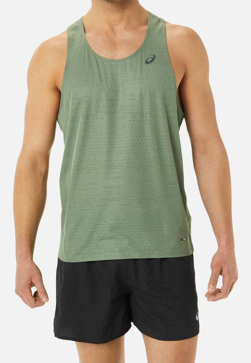 Homme portant un débardeur de sport vert sans manches perforé et un short de sport noir, debout devant un fond uni.