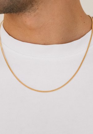 Gul kæde med en rund, klassisk design og en glat overflade, båret over en hvid T-shirt.