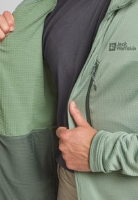 Veste zippée verte avec un tissu texturé et un revêtement lisse, portée par-dessus une chemise noire. Comprend un logo sur la poitrine et une poche zippée.