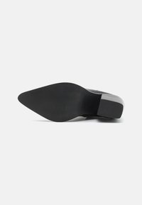 Zapato de tacón negro con punta afilada y suela de goma texturada. Presenta una superficie lisa con un ligero brillo y un tacón cuadrado para mayor soporte.