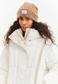 Biała kurtka puchowa z prążkowaną fakturą i beżowymi akcentami, noszona na brązowej, teksturowanej, dzianinowej czapce z naszywką.