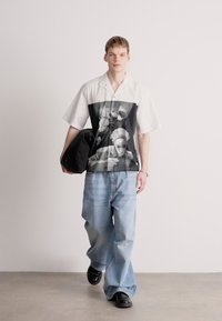 Πουκάμισο κοντό μανίκι με κουμπιά, διακοσμημένο με μαύρη και άσπρη γραφική απεικόνιση στο μπροστινό μέρος, συνδυασμένο με μπιζέ παντελόνια σε χαλαρή γραμμή και μαύρα παπούτσια.