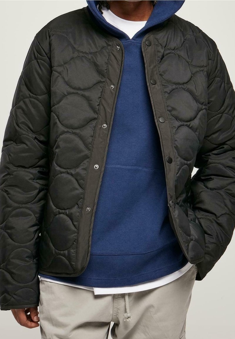 Homme portant une veste matelassée noire sur un sweat à capuche bleu et une chemise blanche avec un pantalon beige, mains dans les poches de la veste, fond neutre.