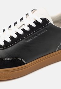 Black leather and suede sneaker with white laces and stitched accents. Features a tan rubber sole and embossed logo detail on the side.