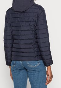 Person, der eine dunkelblaue gesteppte Jacke mit Kapuze und blaue Jeans trägt, von hinten vor einem schlichten Hintergrund gezeigt.