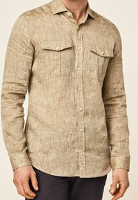 Chemise beige à boutons en tissu texturé, avec deux poches poitrine et manches longues à poignets boutonnés.