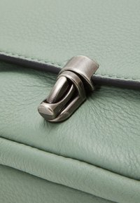 Primer plano de un cierre metálico plateado sobre un material de cuero texturizado verde claro, mostrando las costuras y los bordes doblados de una bolsa o billetera.