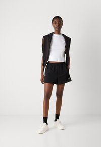 Kvinde i sort ærmeløs top over hvid kort skjorte, sorte shorts, sorte sokker og hvide sneakers stående mod en hvid baggrund.