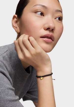 Giovane donna con maglione grigio che appoggia il mento sulla mano, indossando un braccialetto nero intrecciato con un piccolo ciondolo a forma di cuore in oro e un orecchino a cerchio dorato.