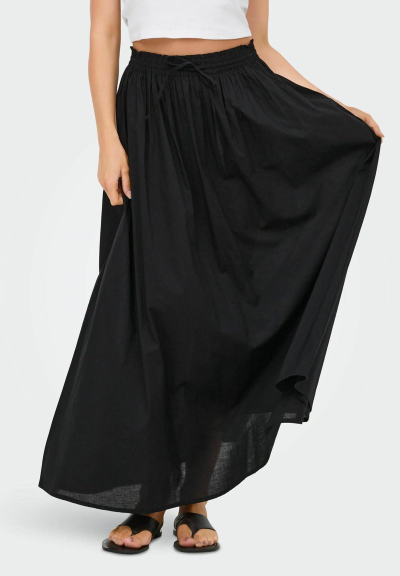 Femme portant une jupe maxi noire fluide avec une taille élastique et un cordon de serrage, accompagnée de sandales noires à bout ouvert sur un fond blanc.