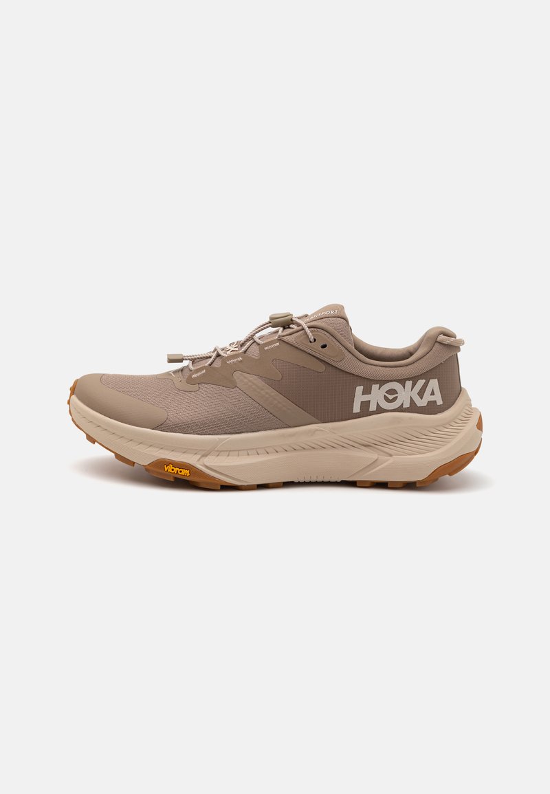 Baskets de course beige clair avec une tige texturée, semelle en caoutchouc et logo HOKA. Présente des lacets élastiques et un talon renforcé pour plus de stabilité.